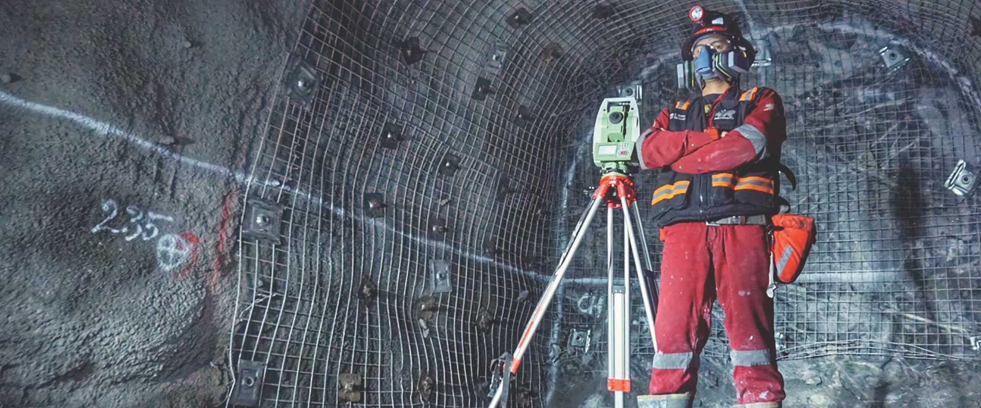 Un employé de Redpath, lourdement vêtu d’équipement de sécurité, y compris un casque, des lunettes et un masque respiratoire, utilise du matériel d’arpentage pour cartographier les angles et les distances dans un tunnel souterrain.