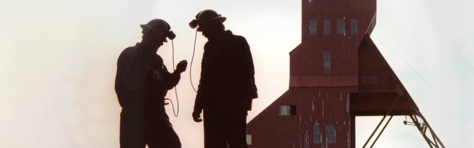 The website banner for the History page shows two silhouetted miners wearing helmets and headlamps, standing on a mine site in front of a weathered mine shaft headframe, engaging in discussion over a device one minor is holding in his hand.