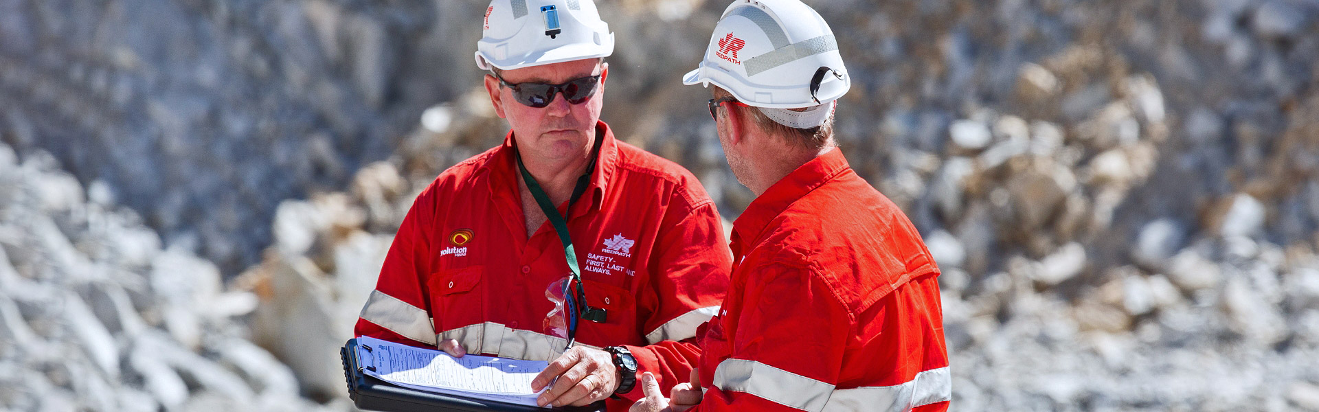 The image shows two Redpath workers, dressed in safety helmets, sunglasses, and bright red coveralls with reflective stripes, standing in an outdoor rocky or quarry-like background, seemingly engaged in a professional discussion or review of documents.