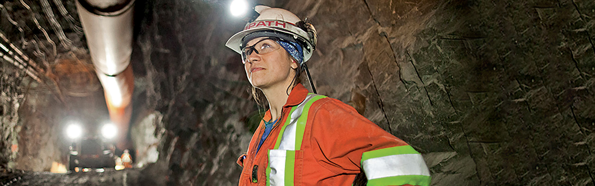 A young female Redpath employee standing in an underground mine tunnel, wearing safety goggles and a hard hat, smiling and looking optimistic.