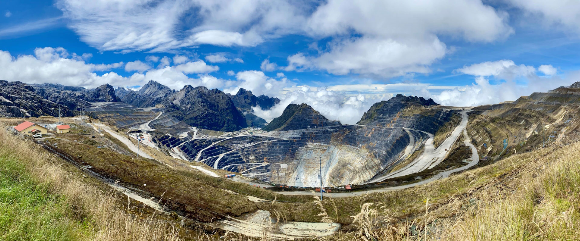 A panoramic photo of a massive Indonesian open-pit mine with layered terrace roads for vehicle access, surrounded by mountains and clouds.