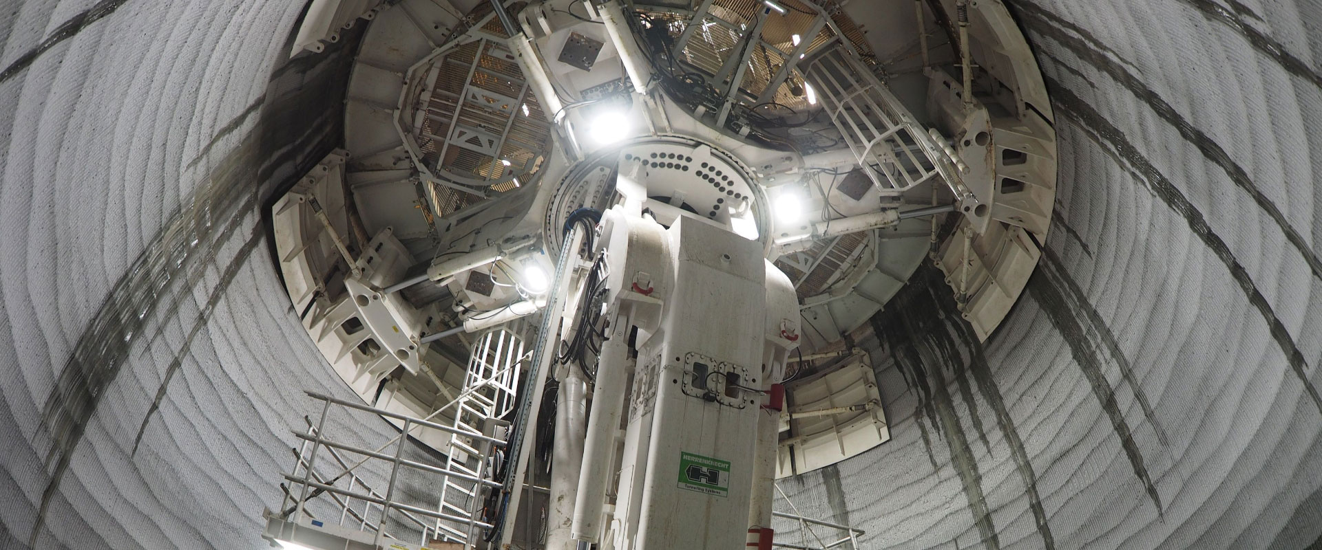A Tunnel Boring Machine (TBM) is a complex piece of engineering, with various components visible: a cutting head or shield at the front, hydraulic supports, and a network of cables and access platforms.